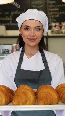Üniformalı kadın aşçı, elinde bir tepsi taze kruvasan tutarken gözleri kapalı gülümser ve fırın mutfağındaki lezzetli aromanın tadını çıkarır..