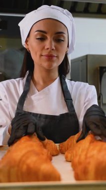 Üniformalı ve eldivenli kadın fırıncı profesyonel mutfak ortamında tepside altın, pullu kruvasanları dikkatlice düzenler..