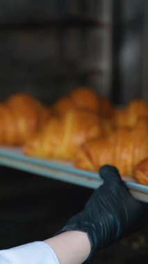 Yeni pişmiş kruvasanların ticari bir mutfakta siyah eldiven giyen bir fırıncı tarafından profesyonel bir fırından çıkarılışının yakın çekim görüntüsü..