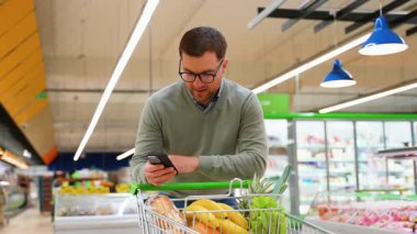 Yeşil kazaklı ve gözlüklü bir adam alışveriş listesini incelerken telefonuna bakar, markette içi taze ürün ve ekmekle dolu bir arabayı iterek..
