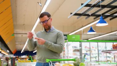 Yeşil kazaklı ve gözlüklü bir adam alışveriş makbuzuna şokla bakar ve yüksek fiyatların farkına varır. Arabası taze ürün ve ekmekle dolu..