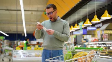 Yeşil kazak ve gözlüklü bir adam, marketteki yüksek fiyatları ve yükselen enflasyonu fark ederek, şok ve inançsızlıkla uzun market faturasına bakar..
