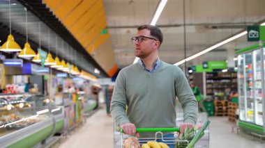 Yeşil kazaklı ve gözlüklü bir adam marketin koridorunda yürüyor, muz, ekmek, ananas ve taze ürünlerle dolu bir arabayı itiyor..