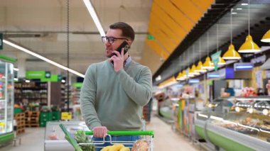 Yeşil kazaklı ve gözlüklü bir adam market koridorunda yürüyor, alışveriş yaparken karısıyla telefonda konuşuyor..