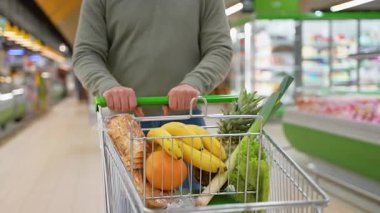Yeşil kazaklı bir adam marketin koridorunda yürürken içi taze meyve, muz, ekmek ve ananas dolu bir alışveriş arabasını itiyor..