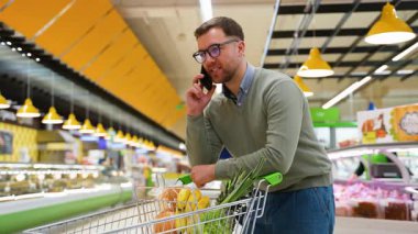 Yeşil kazaklı ve gözlüklü bir adam telefonda konuşurken alışveriş arabasının üzerine eğilir ve karısına süpermarketten ne alması gerektiğini söyler..