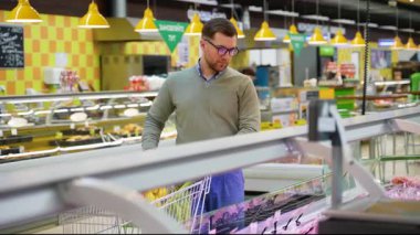 Yeşil süveter ve gözlüklü bir adam süpermarketteki soğutulmuş vitrinden sosis seçiyor, indirimler ve alışveriş yaparken özel teklifler arıyor..