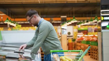 Gözlüklü ve alışveriş arabası olan bir adam dondurucudan donmuş kırmızı balık seçer. O, kaliteli deniz ürünleri bölümünü dikkatle inceliyor..