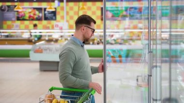 Gözlüklü ve yeşil kazaklı bir adam markette taze somon seçmek için buzdolabının kapısını açar. O kaliteli deniz ürünleri için alışveriş yapıyor..