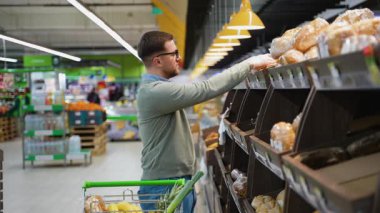 Bir adam süpermarketteki fırın bölümünden taze ekmek toplarken içi alışveriş arabasıyla dolu bir alışveriş arabasını itiyor..