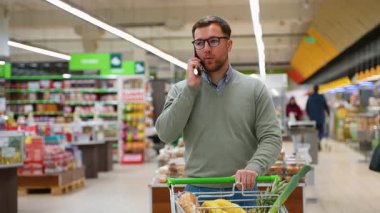 Gözlüklü ve yeşil kazaklı bir adam market koridorunda telefonuyla konuşurken market alışveriş arabasını itiyor..