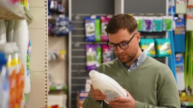 Gözlüklü ve yeşil kazaklı bir adam süpermarket temizlik malzemeleri reyonunda alışveriş yaparken deterjan şişesindeki etiketi dikkatle okuyor..