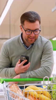 Yeşil kazaklı ve gözlüklü bir adam alışveriş listesini incelerken telefonuna bakar, markette içi taze ürün ve ekmekle dolu bir arabayı iterek..