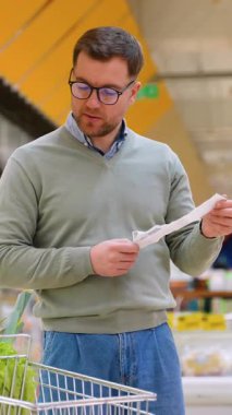 Yeşil kazaklı bir adam, alışveriş arabasının yanında durup, süpermarketteki yeni ürünlerin masraflarını hesaplarken, uzun bir market fişini inceliyor..