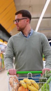 Yeşil kazaklı ve gözlüklü bir adam marketin koridorunda yürüyor, muz, ekmek, ananas ve taze ürünlerle dolu bir arabayı itiyor..