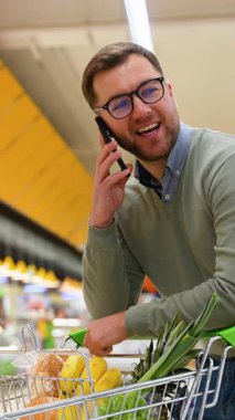 Yeşil kazaklı ve gözlüklü bir adam telefonda konuşurken alışveriş arabasının üzerine eğilir ve karısına süpermarketten ne alması gerektiğini söyler..