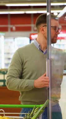 Yeşil kazaklı ve gözlüklü bir adam süpermarkette soğutmalı vitrini açıyor, alışveriş arabasıyla alışveriş yaparken indirimli jamonu seçiyor..
