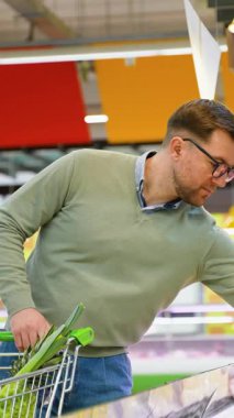 Yeşil kazaklı bir adam süpermarketteki soğutuculu bölümden dikkatlice peynir seçerken bir alışveriş arabasını taze yiyeceklerle itiyor..