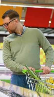 Yeşil kazaklı bir adam buzdolabı bölümünden gurme peyniri seçerken içi taze ürünlerle dolu bir alışveriş arabasını itekliyor..