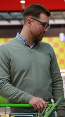 Yeşil kazaklı bir adam içecek reyonuna bakıyor, karbonatlı içecekleri dikkatlice seçerken içi taze yiyecek dolu bir alışveriş arabasını itiyor..