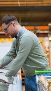 Gözlüklü ve alışveriş arabası olan bir adam dondurucudan donmuş kırmızı balık seçer. O, kaliteli deniz ürünleri bölümünü dikkatle inceliyor..