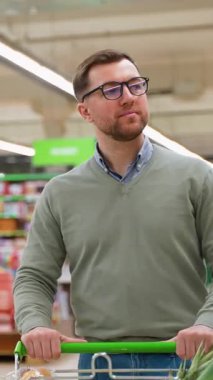 Yeşil kazaklı ve gözlüklü bir adam, marketin taze ürünler reyonuna bakarken muz, ananas, marul ve ekmekle dolu bir alışveriş arabasını itiyor..
