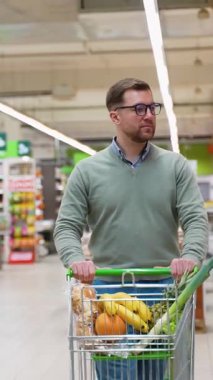 Gözlüklü bir adam ve süpermarkette yeşil bir kazak dükkanı, içi taze ürün, ekmek ve diğer yiyecek malzemeleriyle dolu bir market arabasını itiyor..