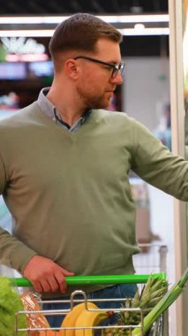 Yeşil kazaklı ve gözlüklü bir adam marketin sebze reyonunda taze salata seçeneklerine bakmış ve sağlıklı bir seçim yapmış..