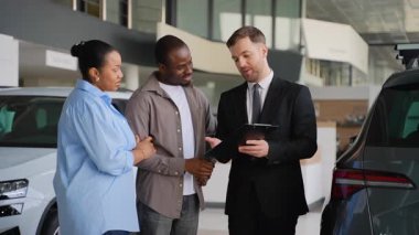Profesyonel otomotiv danışmanı, Afro-Amerikan çiftlere araç seçimi sürecinde rehberlik ediyor, lüks modern galeri galerisindeki seçenekleri yönlendiriyor.