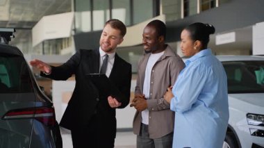 Afro-Amerikan bir çifte yeni bir araba sunan profesyonel bir araba satıcısı, modern bir galeride elinde bir pano tutarken özellikleri ve faydaları açıklıyor.