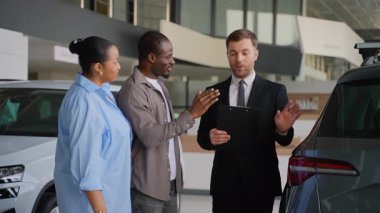 Profesyonel satış danışmanı, modern bayilik galerisinde araba seçimi sürecinde Afro-Amerikan çiftlere rehberlik ederek, kişisel otomotiv satın alma tavsiyeleri sunuyor.