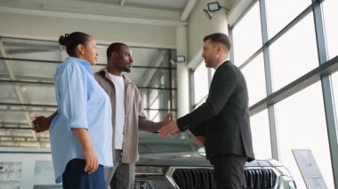 Profesyonel araba satıcısı, Afro-Amerikan çifti karşılıyor, el sıkışıyor ve modern, parlak galeride başarılı araç alımlarını kutluyor.