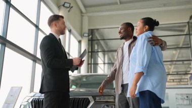 Satış danışmanı heyecanlı Afro-Amerikan çifti için lüks bir araç sunuyor, anahtar özellikleri açıklıyor ve çağdaş bayilikte profesyonel el sıkışma anlaşmasını mühürlüyor.