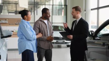 Heyecanlı Afro-Amerikan çifti yeni araba anahtarlarını galerideki profesyonel bir araba satıcısından alıyor. Satışlarını neşe ve coşkuyla kutluyorlar.
