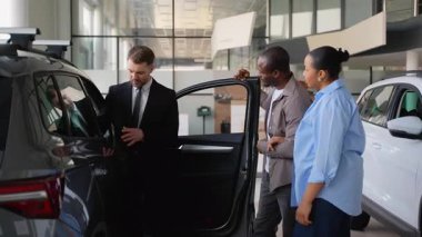 Afro-Amerikan bir çifte yeni bir arabanın özelliklerini gösteren profesyonel bir araba satıcısı modern bir bayilikte doğru seçimi yapmalarına yardım ediyor.