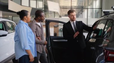 Afro-Amerikalı çift, satış mağazasındaki yeni aracı dikkatle inceliyor. Detayları tartışırken ve otomotiv seçeneklerini incelerken dikkatli pazarlamacılarla profesyonel etkileşimde bulunuyor.
