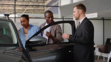 Profesyonel araba satıcısı, Afrikalı Amerikalı çifte yeni araba özelliklerini gösteriyor. Modern galeride bilinçli karar vermelerine yardımcı oluyor.