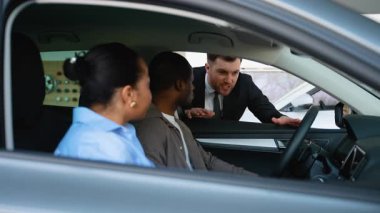 Deneyimli bir araba satıcısı, modern galeride yeni araç özelliklerini incelerken Afrikalı Amerikalı bir çifte detaylı rehberlik ediyor.