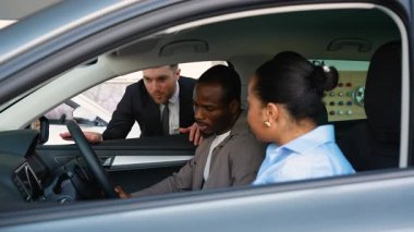 Profesyonel araba satıcısı Afro-Amerikan çiftlere rehberlik ederken modern galeride araç özellikleri seçiyor, kişisel otomotiv satış deneyimleri sergiliyor.