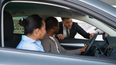 Araba satıcısı, araba seçimi sürecinde Afro-Amerikan çiftlere rehberlik ediyor, galeri galerisinde profesyonel uzmanlık sağlarken araç özelliklerini ve seçeneklerini araştırıyor.