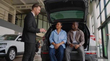 Açık bagajda oturan Afro-Amerikan çifte bagajlarını gösteren profesyonel bir araba satıcısı modern galeride yeni bir aracın özelliklerini ve faydalarını tartışıyor.