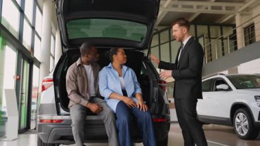 Araba satıcısı, modern galeride potansiyel araç alımını değerlendiren Afrikalı Amerikalı çifte geniş bir sandık alanı gösteriyor.