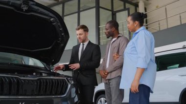 Profesyonel araba satıcısı, modern galeride otomotiv satış danışmanlığı sırasında motor özelliklerini vurgulayan Afrikalı Amerikalı çifte araç detaylarını sunuyor.