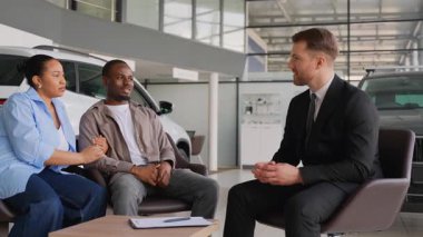 Profesyonel araba satıcısı araç seçeneklerini sunuyor, modern galeri galerisinde detaylı satış danışmanlığı sırasında Afrikalı Amerikalı çifti meşgul ediyor.
