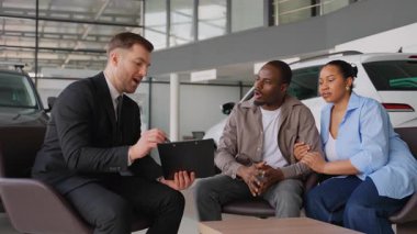 Profesyonel satış danışmanı, Afro-Amerikan çiftlere araba seçimi sürecinde rehberlik ediyor, modern galeride araç detaylarını tablete aktarıyor.