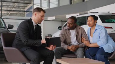Profesyonel bir araba satıcısı, yeni arabalarla çevrili bir oto galerisindeki koltukta oturan Afro-Amerikan bir çifte kontratı gösteriyor ve açıklıyor.