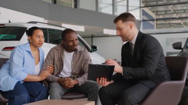 Satış danışmanı, Afro-Amerikan çiftlere modern bayilik galerisinde araba satın alma sürecinde rehberlik ederken, araç seçimini ve alımını kolaylaştırıyor.
