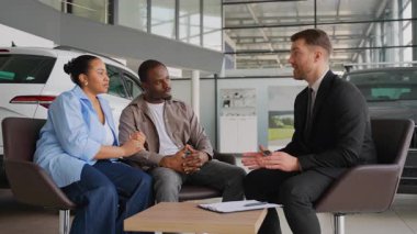 Profesyonel araba satıcısı heyecanlı bir Afro-Amerikan çifti, lüks, modern otomobil galerisinde araç seçimi sürecinde, satın alma detaylarını tartışıyor ve finansman seçeneklerini araştırıyor.