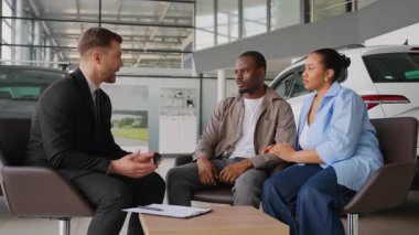 Takım elbiseli satıcı, modern araba galerisindeki koltukta oturan neşeli Afro-Amerikan çifte araba anahtarlarını veriyor. Başarılı bir satın alımı kutluyorlar.