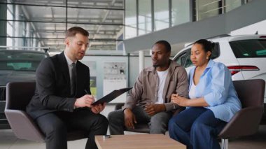 Afro-Amerikan çifte belgeleri gösteren profesyonel bir araba satıcısı, galeride yeni bir araba seçip almalarına yardım ediyor.